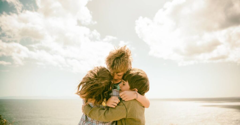 three children hugging at the beach in front of the ocean as they offer each other Christian forgiveness